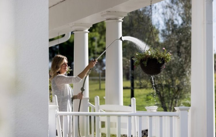 Woman watering a hanging flower basket with a Hoselink Long Reach Shower Wand on a sunny porch with white railing and columns