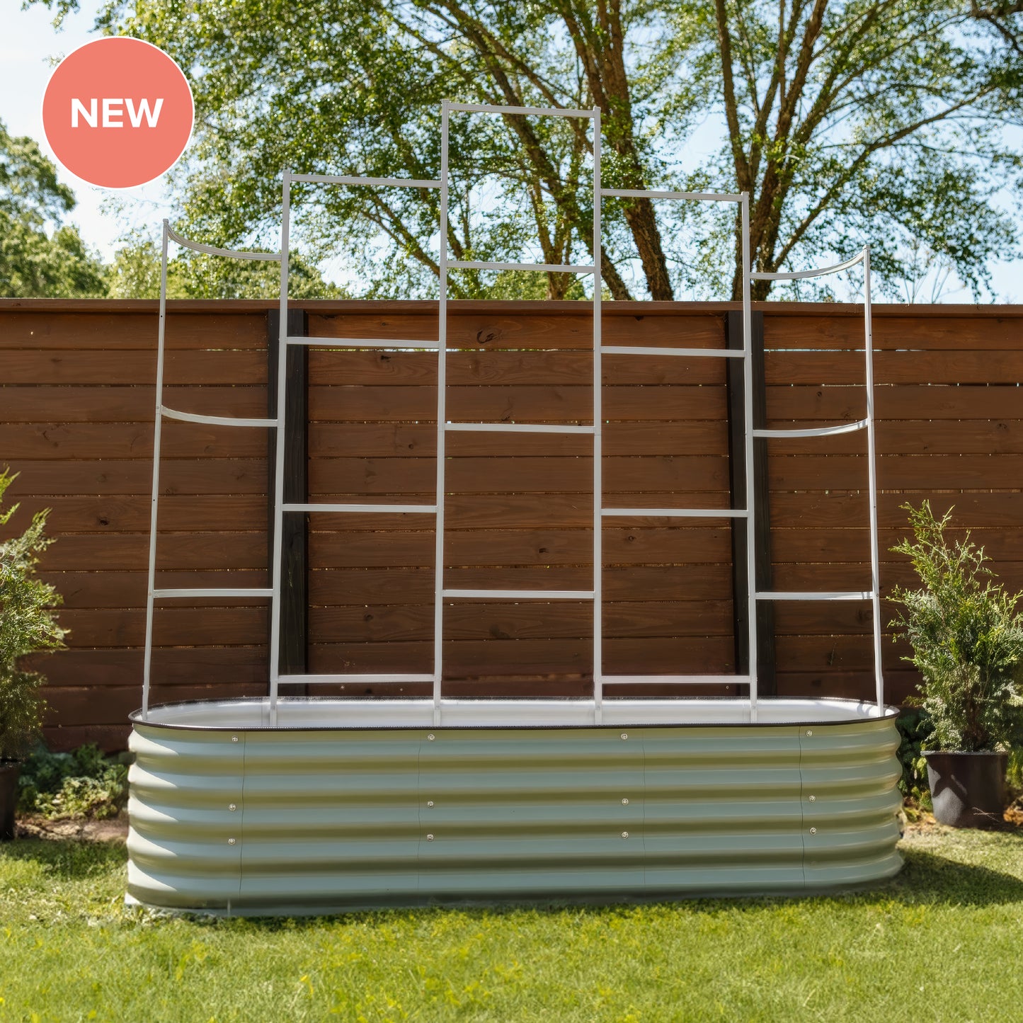 A corrugated metal garden bed with a white metal ladder-style trellis frame set up outdoors on grass in front of a wooden fence. A pink circular 'NEW' badge appears in the top-left corner.