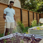 Man tending to plants in a garden with a hose and a wooden fence and trees in the background