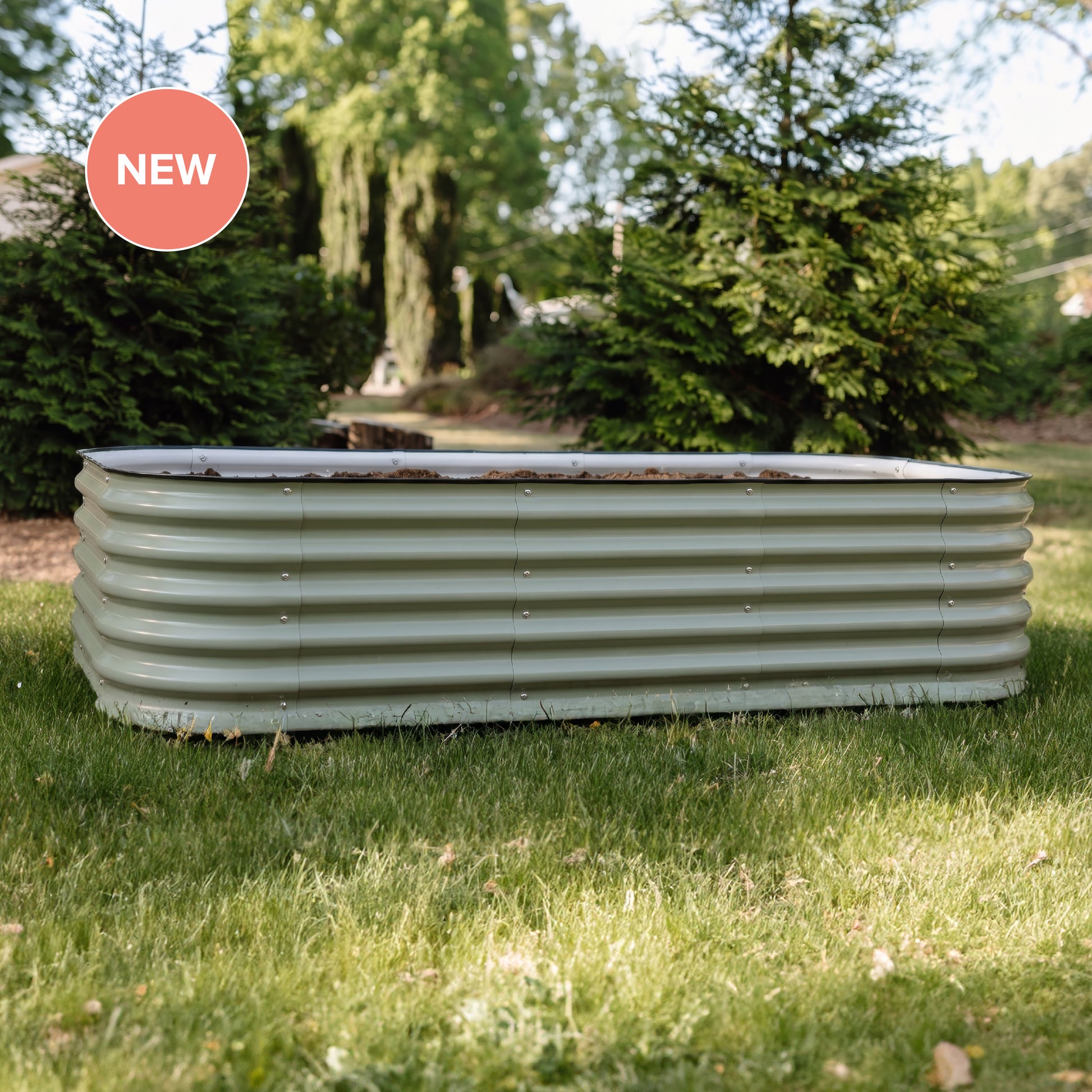 A rectangular corrugated metal garden bed in a sage green color, placed on a grassy lawn with trees in the background. A pink circular 'NEW' badge is in the top left corner.