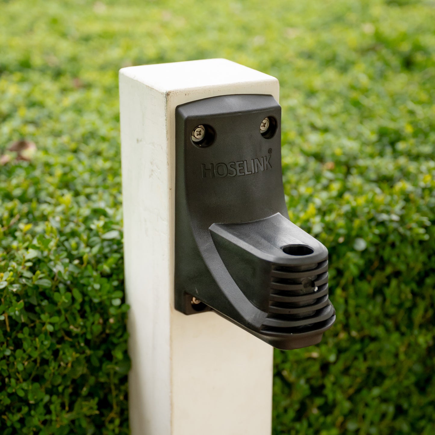 Close up of a hose reel wall bracket installed on a white post, in front of a hedge.