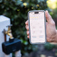 Hand holding a smartphone with a smart water timer app displayed, with a garden faucet and dual outlet timer in the background.