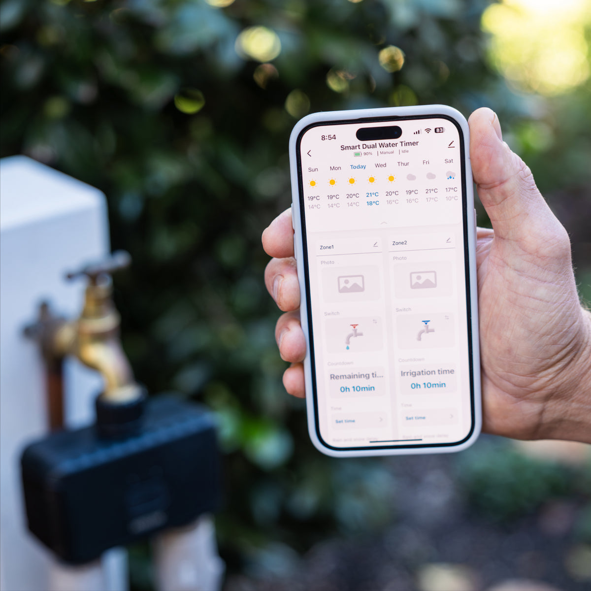 Hand holding a smartphone with a smart water timer app displayed, with a garden faucet and dual outlet timer in the background.