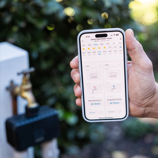 Hand holding a smartphone with a smart water timer app displayed, with a garden faucet and dual outlet timer in the background.