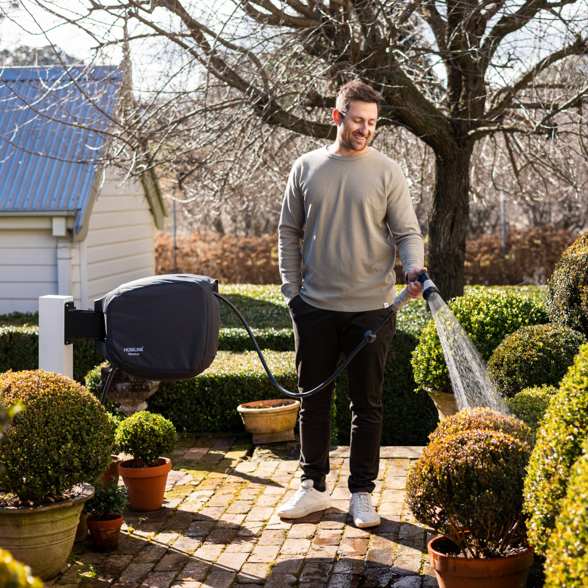 Man watering plants in a garden with an evolve hose reel with charcoal cover, surrounded by potted plants and a house in the background.