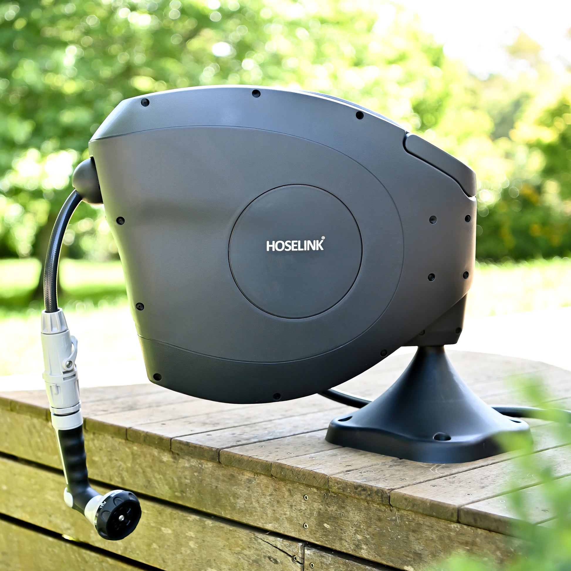 A Hoselink retractable reel mounted on a fixed ground mount on a timber deck, with greenery in the background.