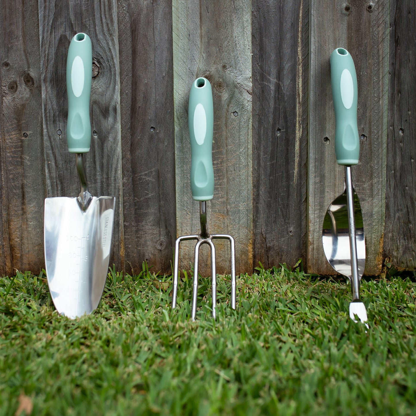 Garden tool set with green handles on grass against a wooden fence.