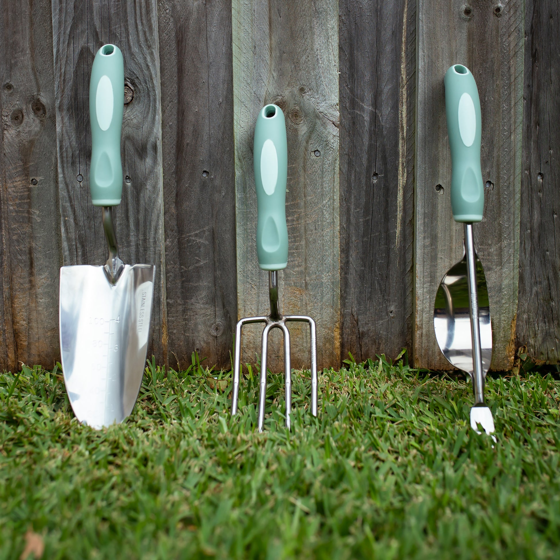 Garden tool set with green handles on grass against a wooden fence.