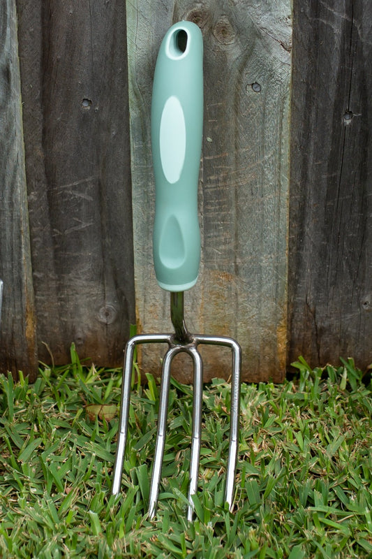 A garden hand fork with a mint green and white ergonomic handle and silver metal tines, standing upright in green grass against a weathered wooden fence.