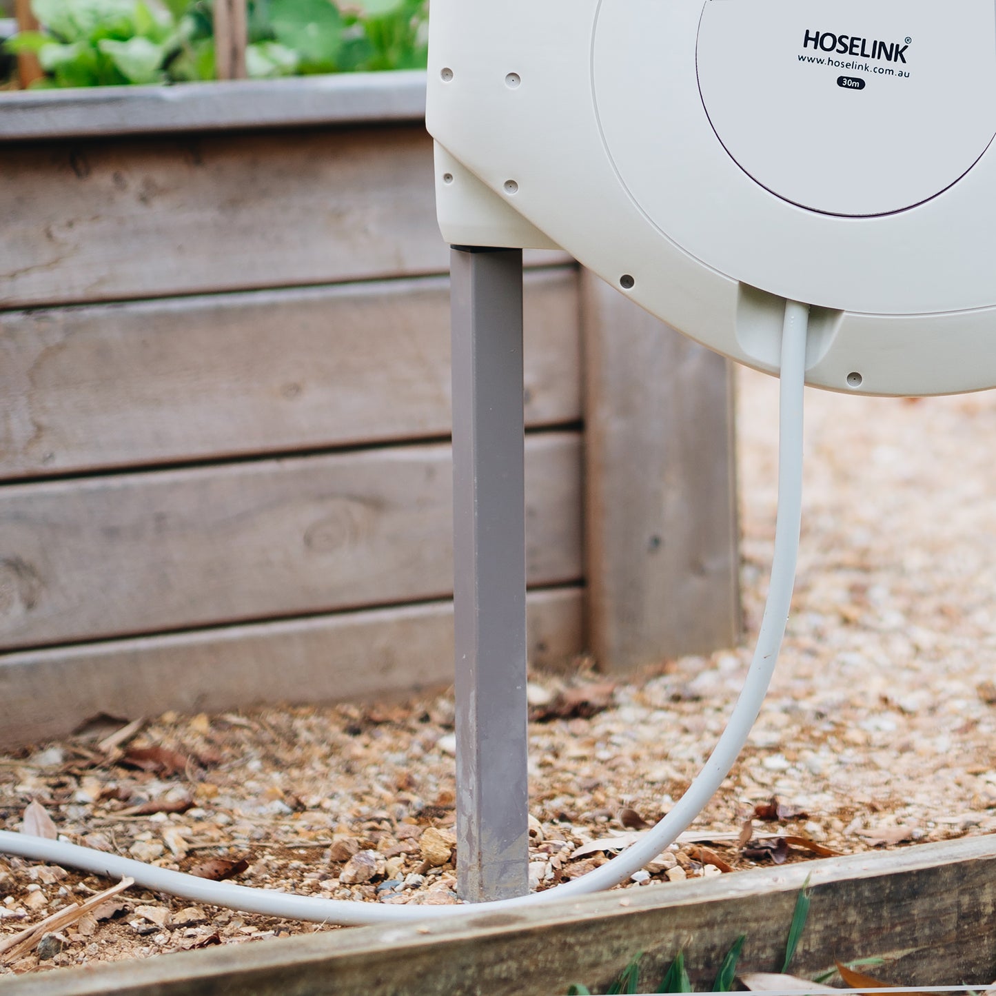 Beige retractable hose reel mounted onto a grey powdered coated stainless steel post in a garden bed