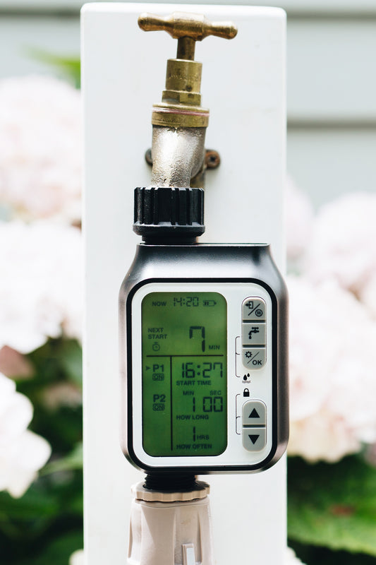 A timer with a digital LCD display and five buttons connected to a garden faucet with hose connector underneath and pale pink flowers in the background.