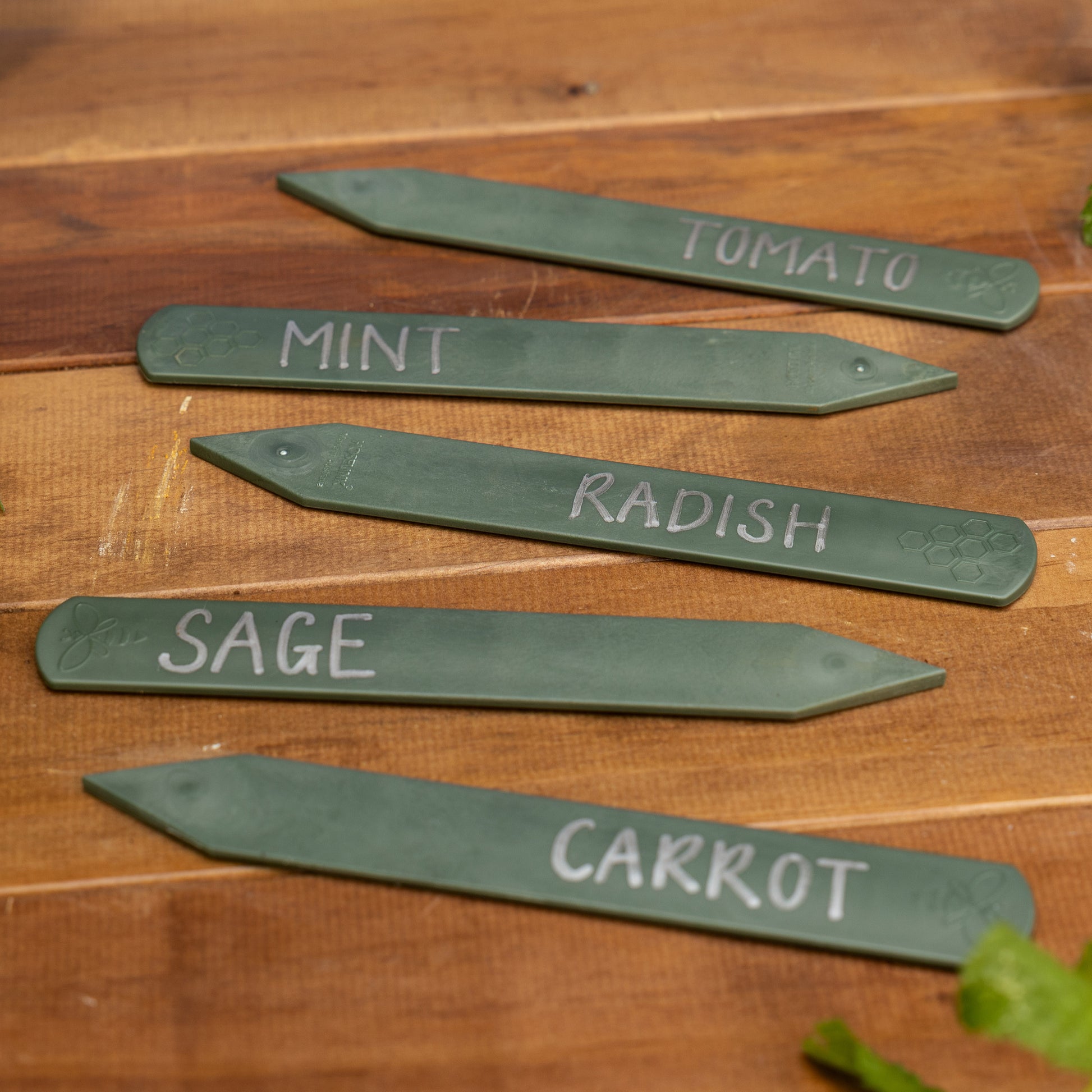 Set of five recycled plastic plant labels arranged on a wooden bench with different herbs and vegetables written on