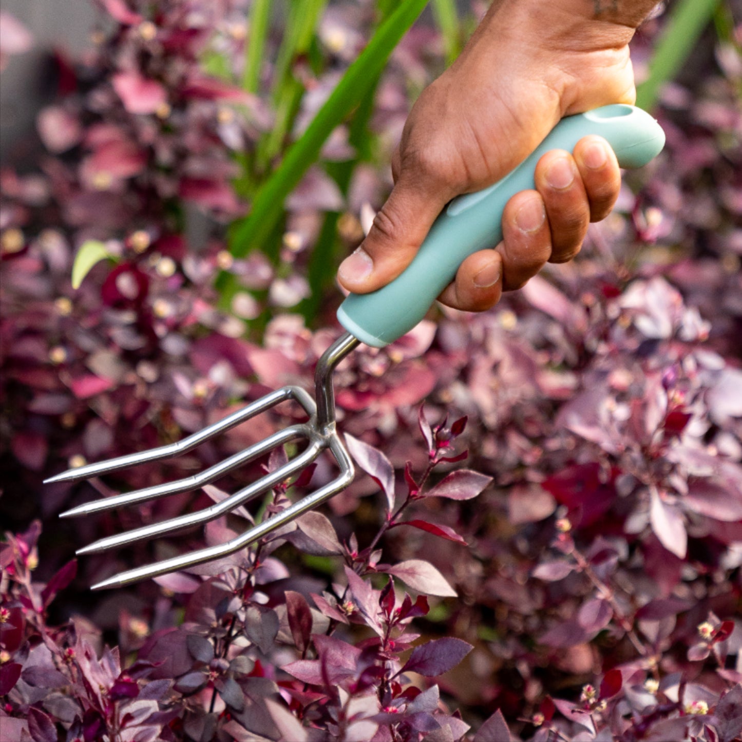 A hand holding the Garden Fork in a garden setting with plants and a wooden fence.