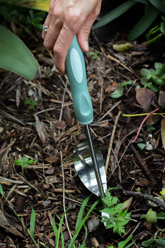 A close-up of a hand holding a small garden hand tool with a sage green handle and a stainless steel blade, digging into mulch and soil outdoors.