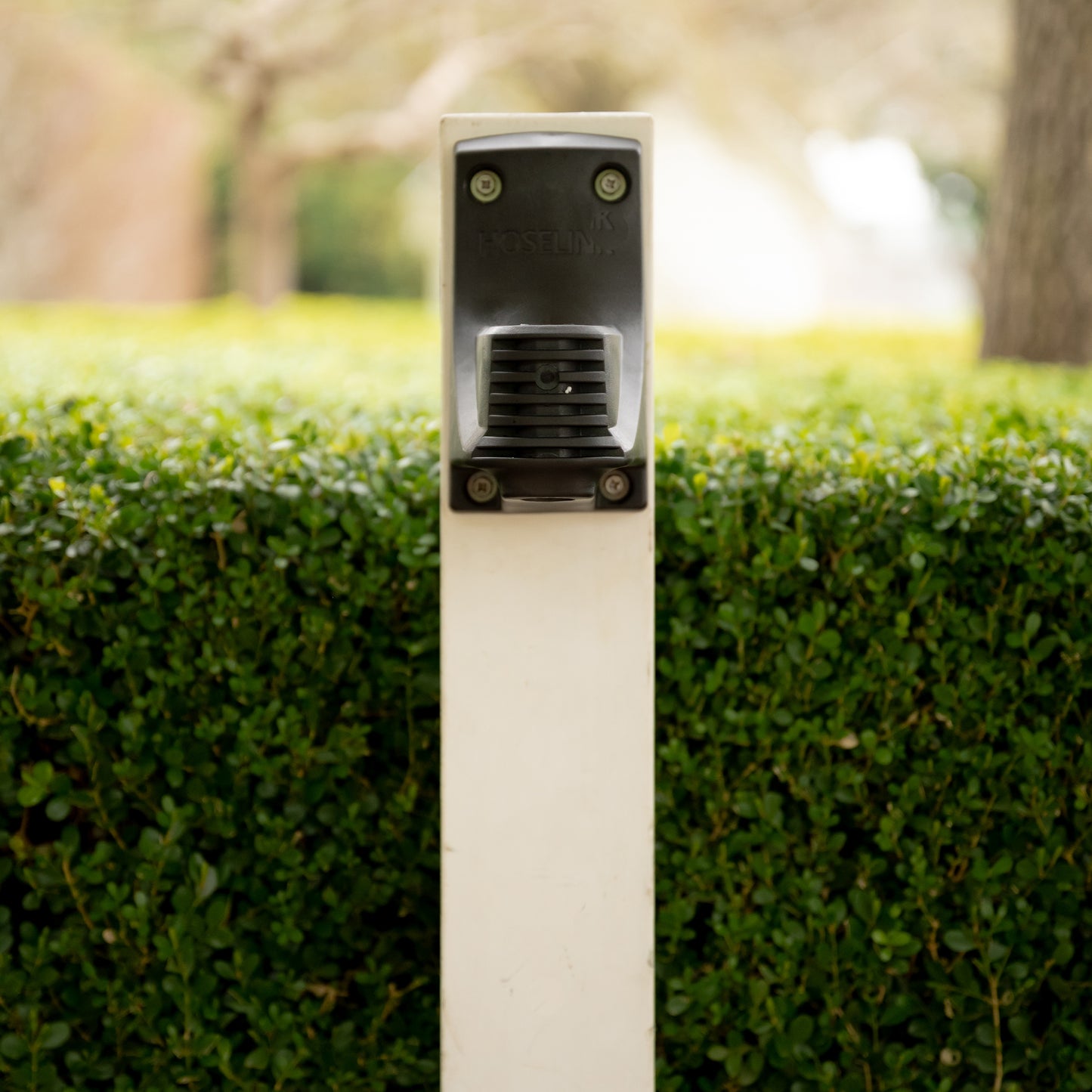 A hose reel wall bracket installed on a white post, in front of a hedge.