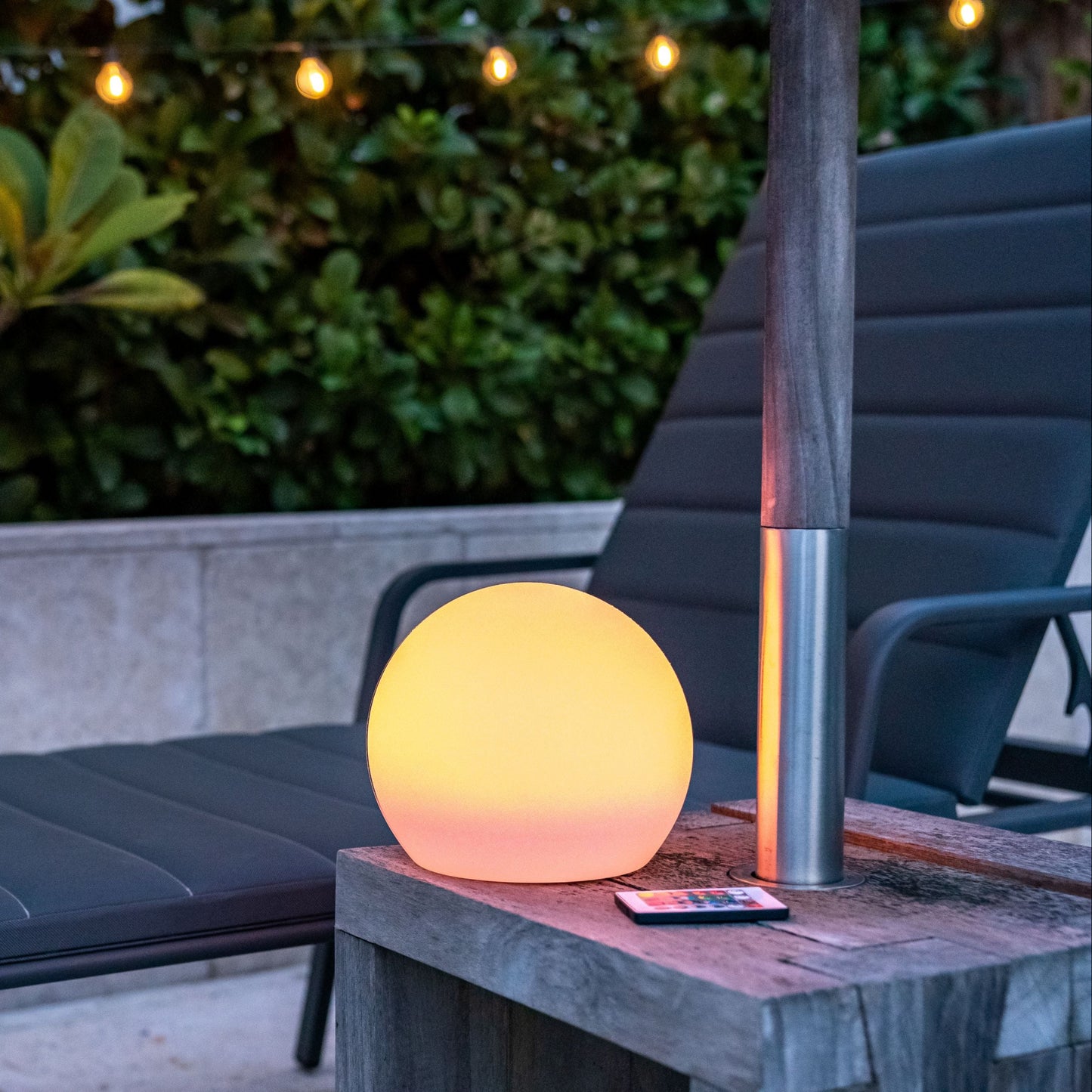 Outdoor setting with a solar sphere light on a table, surrounded by greenery and a lounge chair.