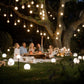 People enjoying a picnic under a tree with hanging lights and solar sphere lights at night.