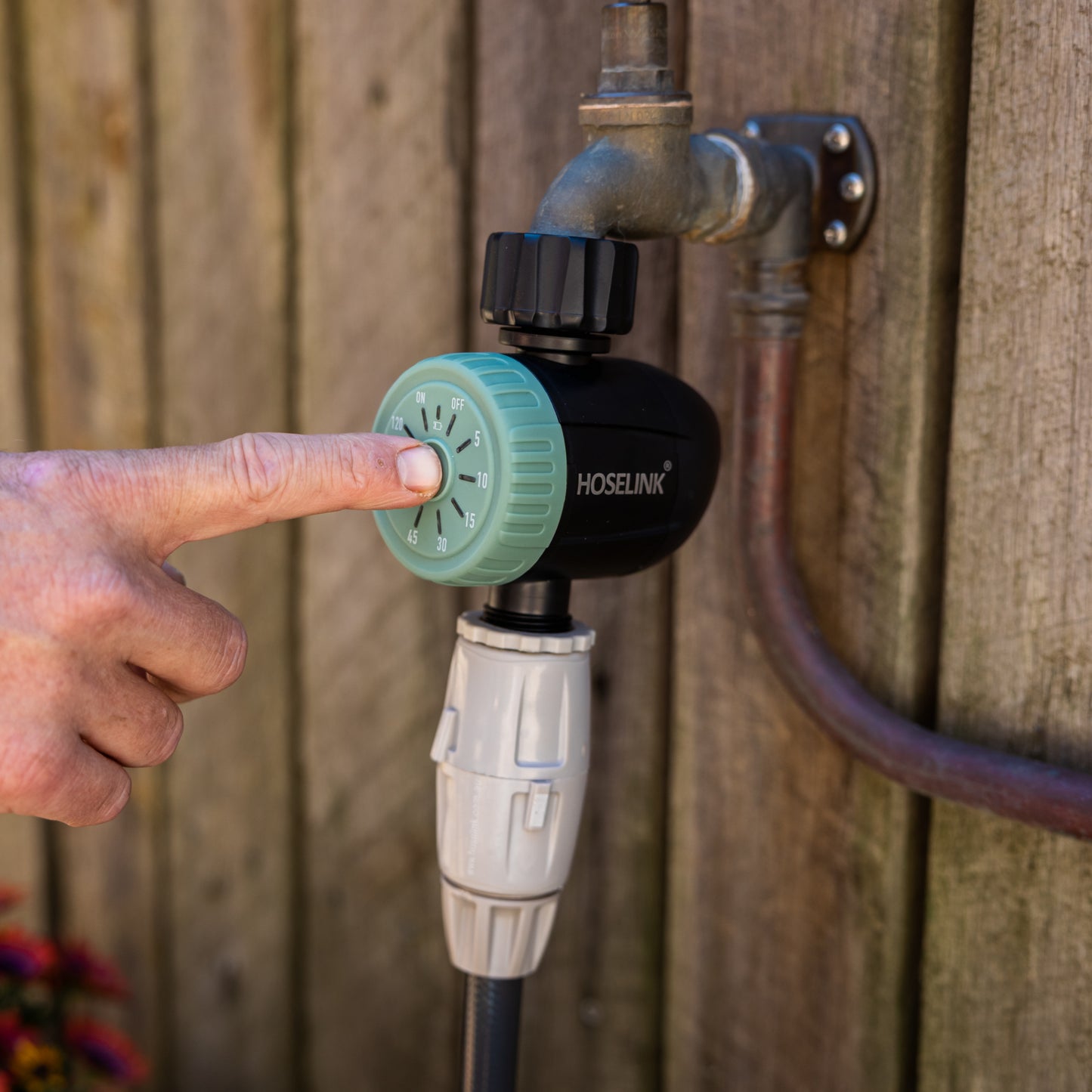 Hand adjusting a green dial timer on a garden faucet against a wooden fence background