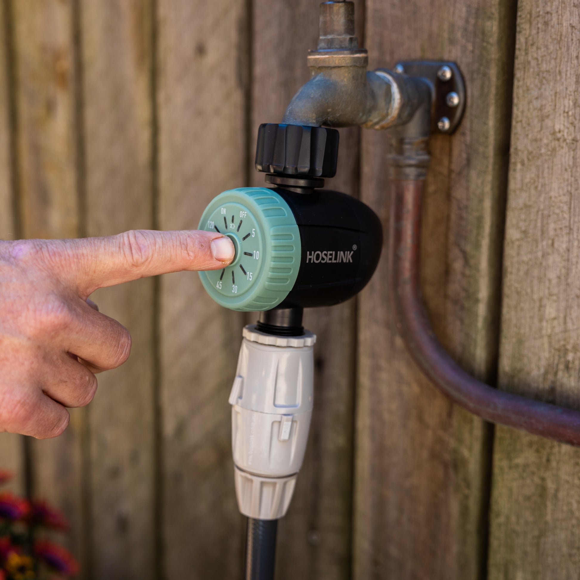 Hand adjusting a green dial timer on a garden faucet against a wooden fence background