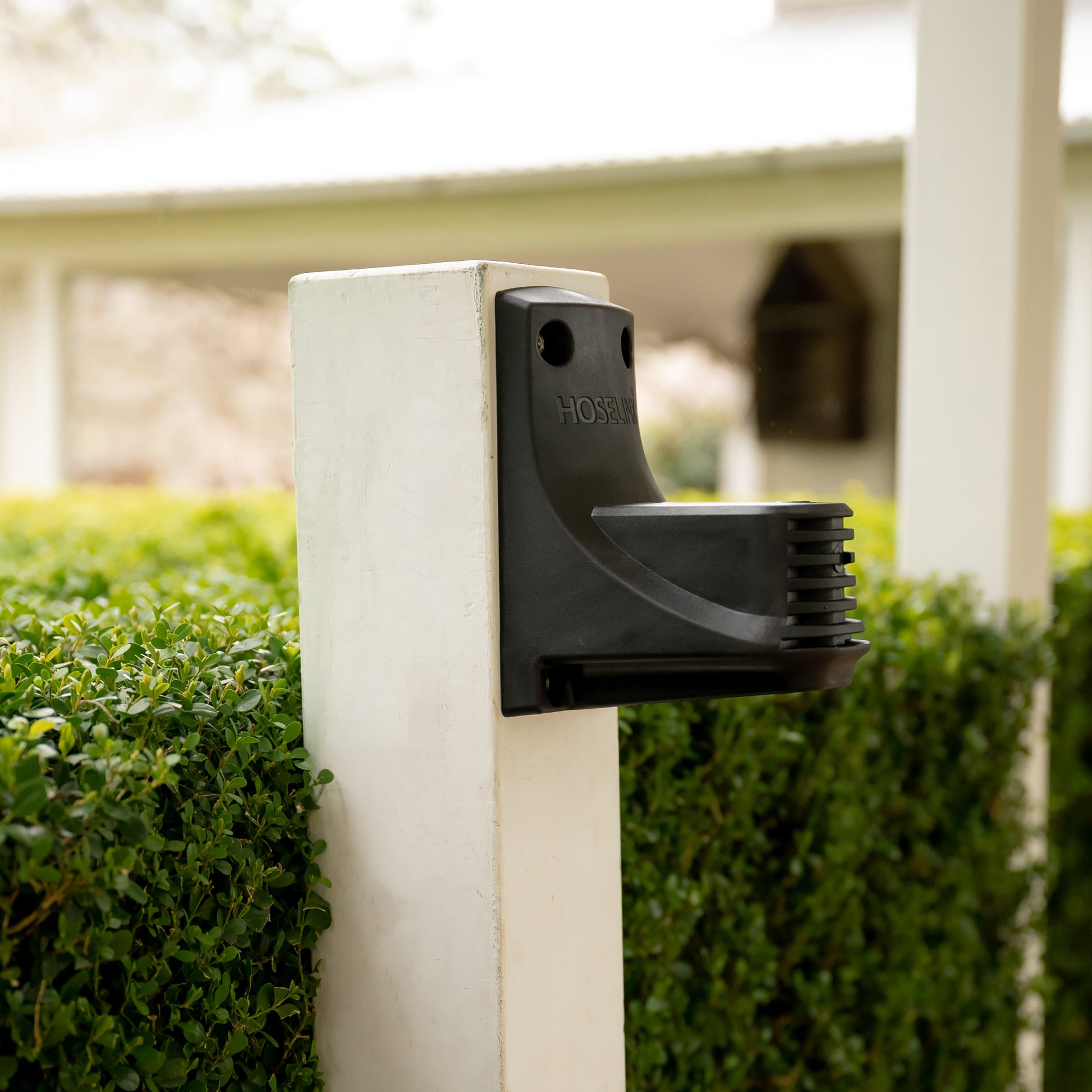 Close up of the rear of the hose reel wall bracket installed on a white post, in front of a hedge.