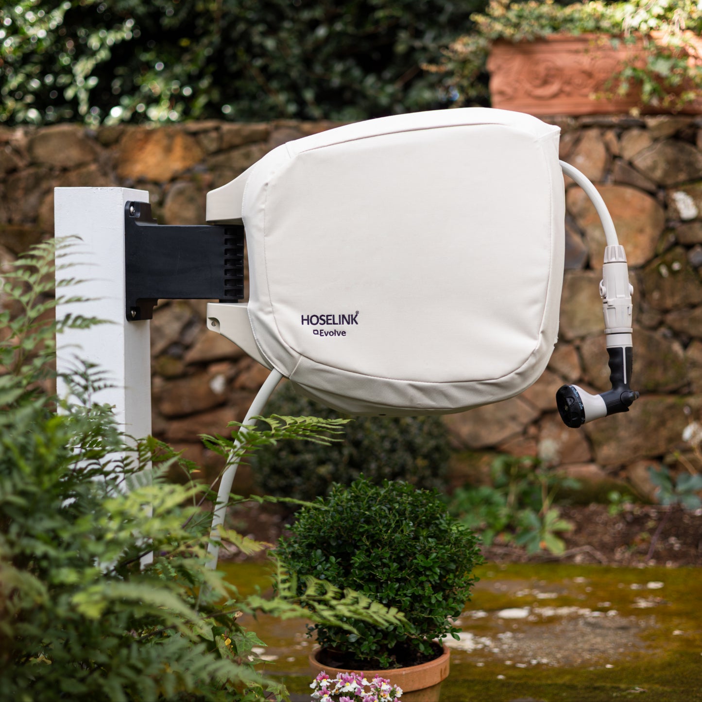Beige hoselink Evolve hose reel cover on hose reel, mounted outdoors with plants and a stone wall in the background.