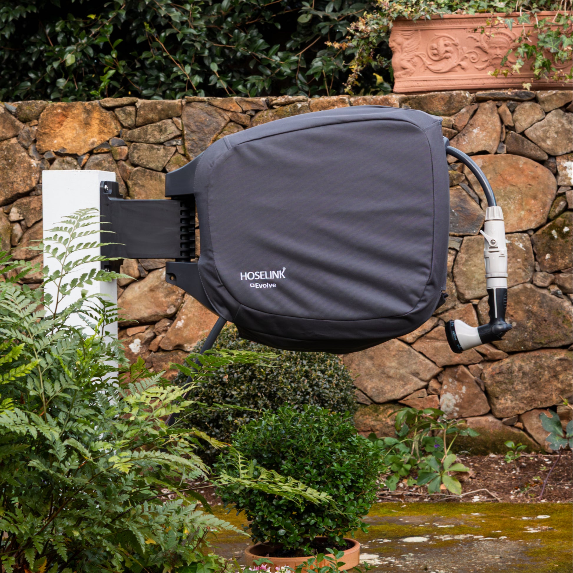 hoselink evolve reel with charcoal cover mounted on a white post with plants and stone wall in the background.