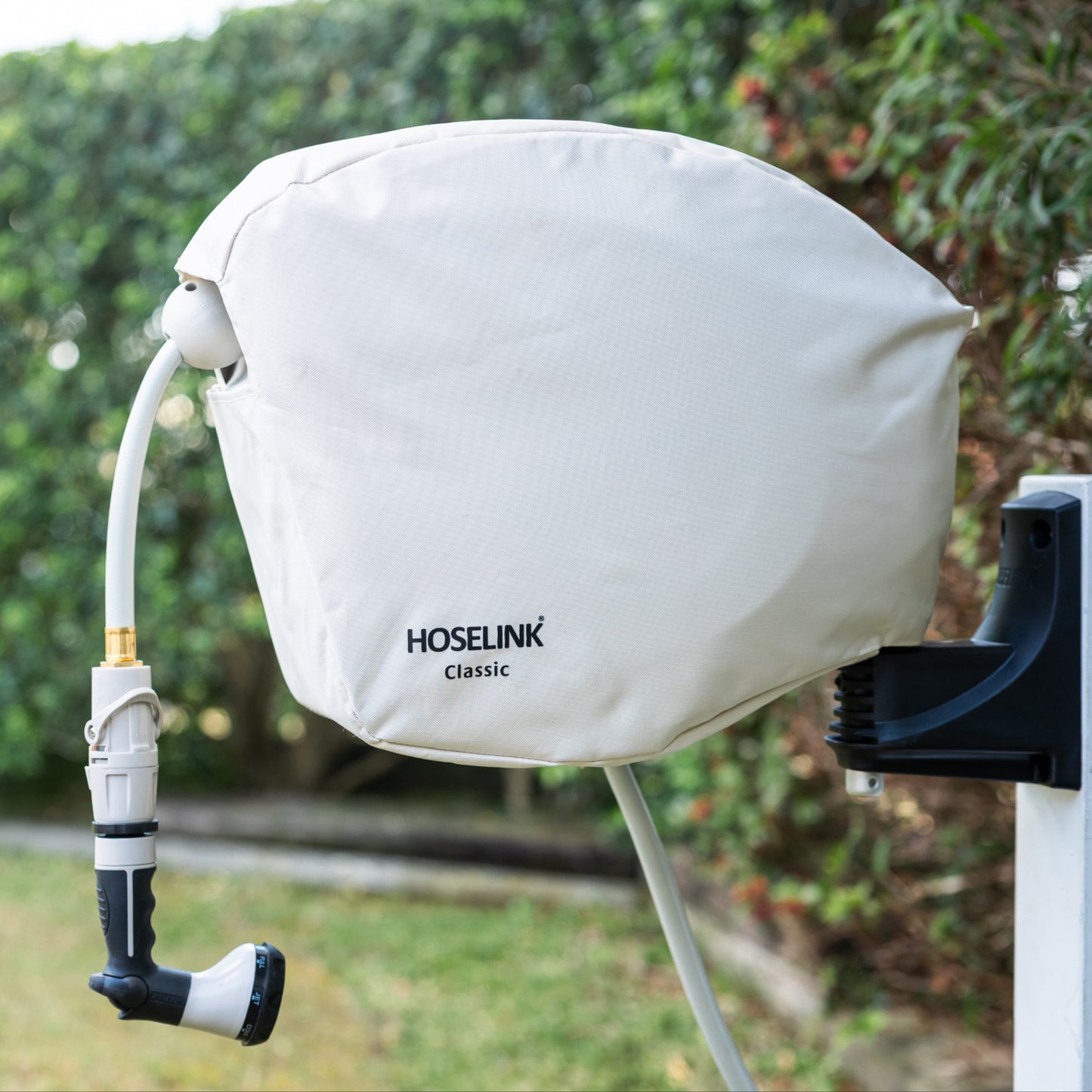 Beige classic hose reel cover with black Hoselink logo on a hose reel in a backyard with green lawn and hedges in the background