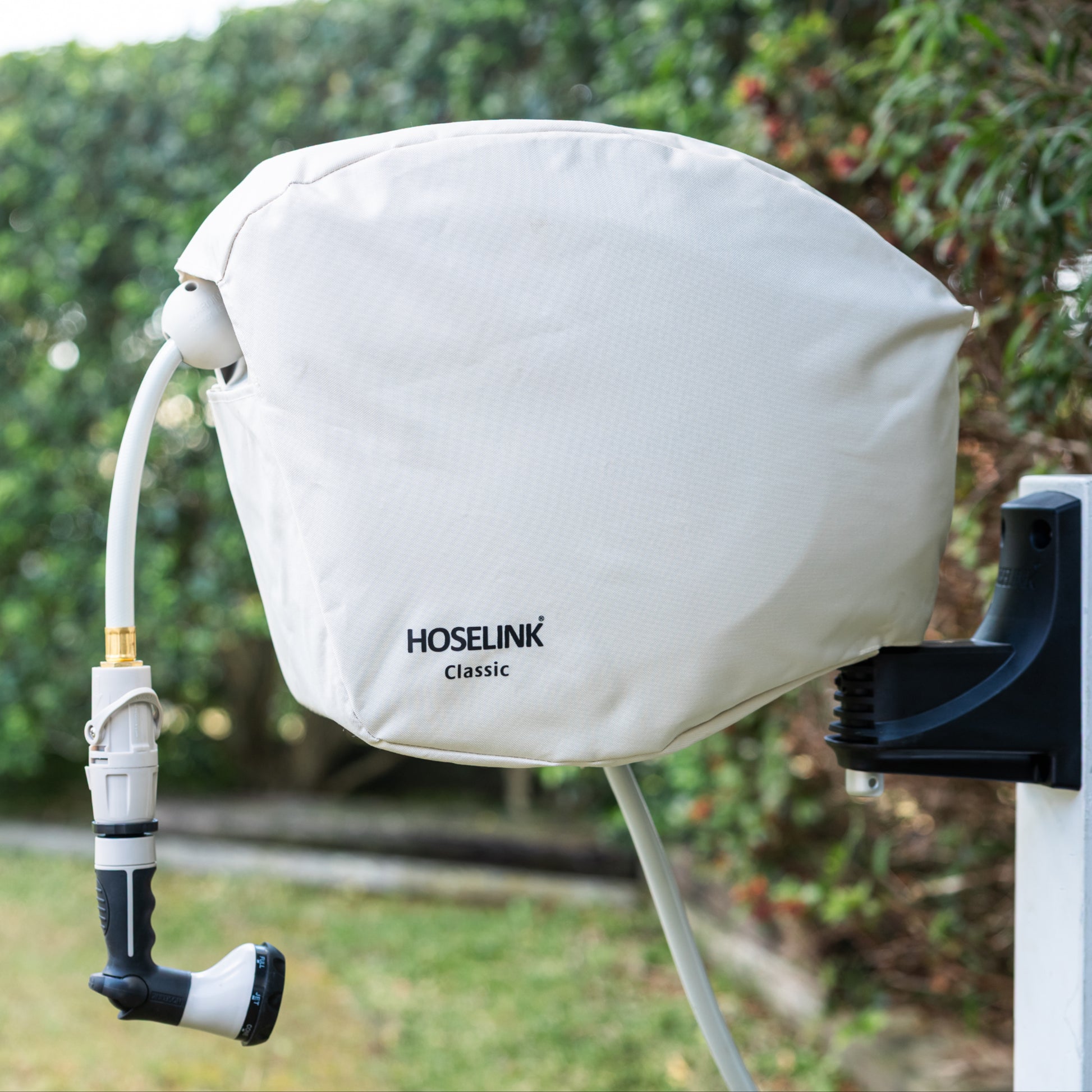 Beige classic hose reel cover with black Hoselink logo on a hose reel in a backyard with green lawn and hedges in the background