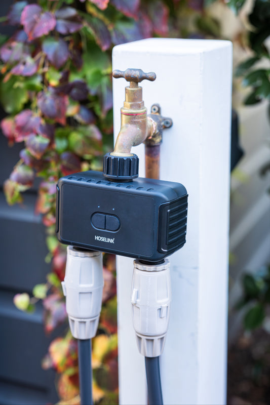 smart hose time dul outlet installed on an outdoor faucet on a white post with two hoses attahced and vines in the background