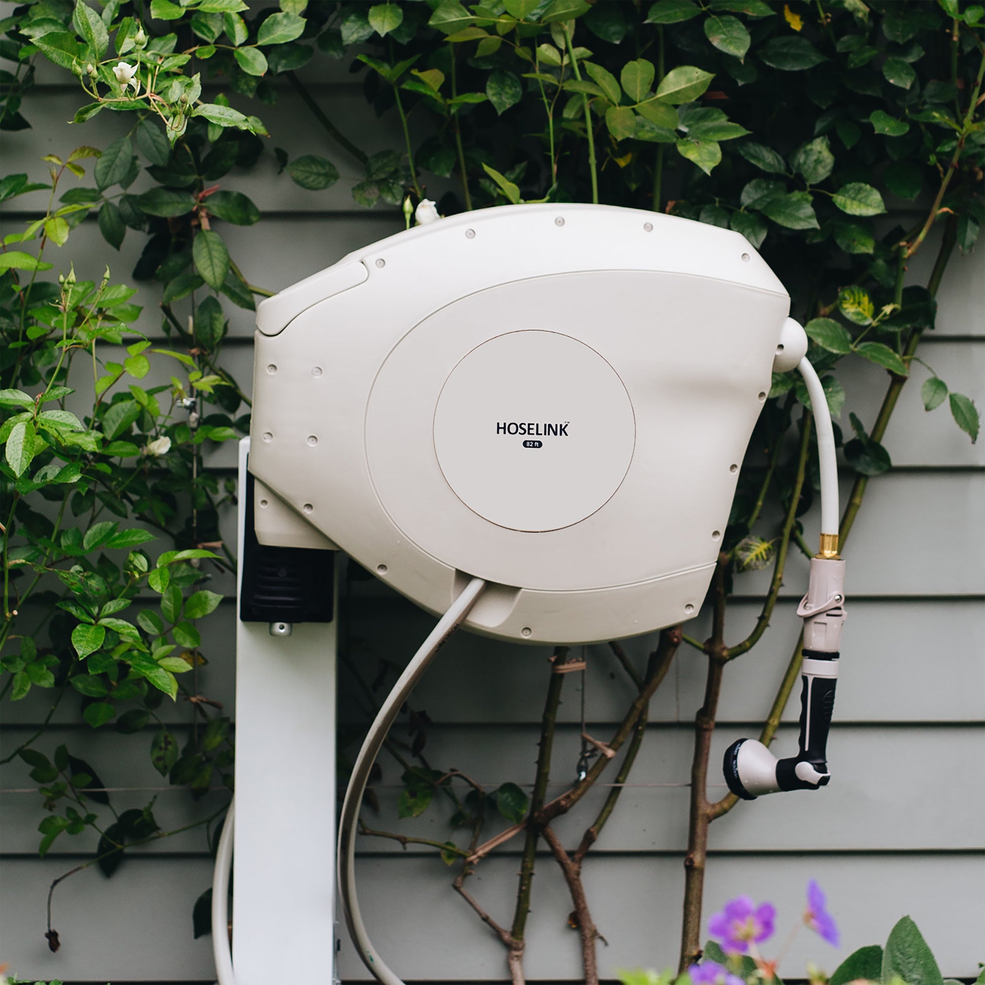 beige hose reel mounted on a post against a grey weatherboard house with a climbing plant 