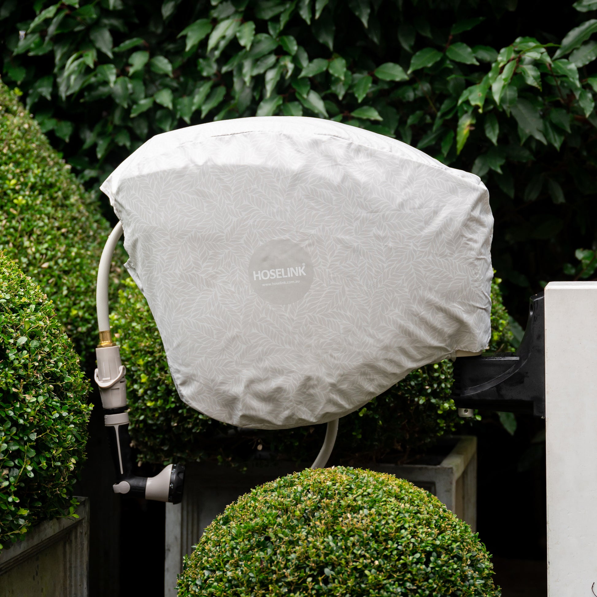 beige hose reel mounted on a post amongst hedges with a beige fern hose reel cover over it